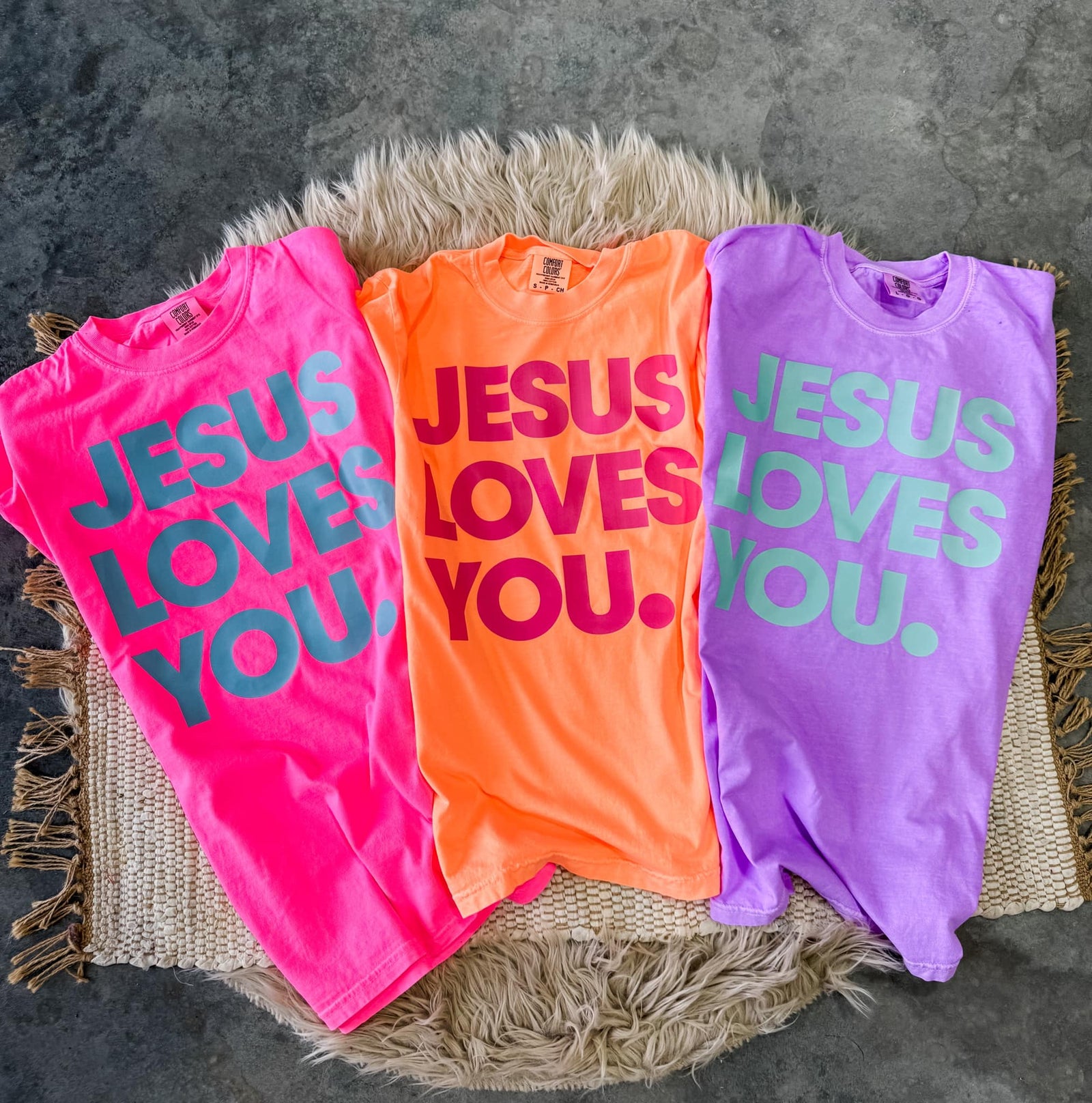 Three t-shirts in pink, orange, and purple colors laid out flat with the text 'JESUS LOVES YOU' printed in bold letters.